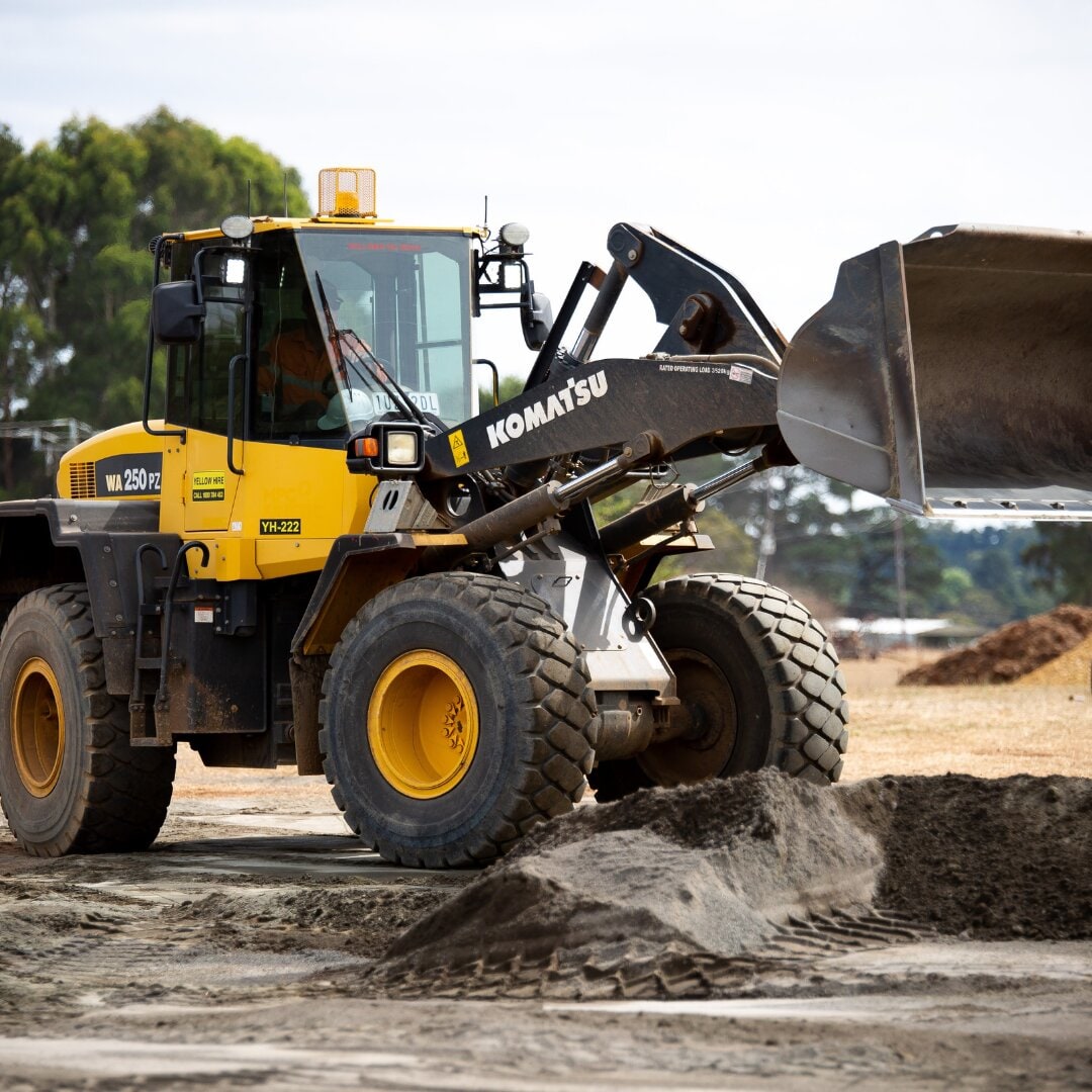 A Komatsu WA250 wheel loader scooping a load of dark soil from a pile on a job site.