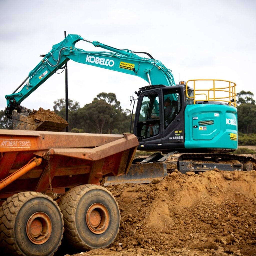 A teal Kobelco SK135SR excavator swinging its arm to dump a load of dirt into a large orange haul truck.